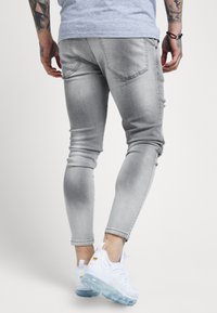 Jeans de mezclilla gris de corte slim con un ligero desgaste y detalles desgastados, combinados con zapatillas blancas con suela transparente.