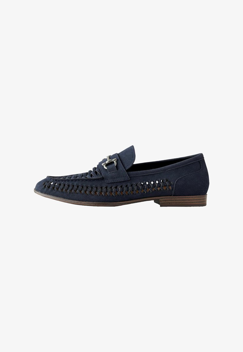 Mocassin en daim bleu marine avec des détails tissés sur les côtés, une bride à boucle argentée et un petit talon en bois sur fond blanc.