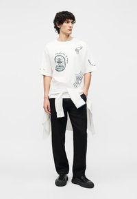 Νέος άντρας με κατσαρά μαλλιά που φοράει λευκό μπλουζάκι με σχέδιο, μαύρο παντελόνι, μαύρα αθλητικά παπούτσια και ένα λευκό πουκάμισο δεμένο γύρω από τη μέση του.