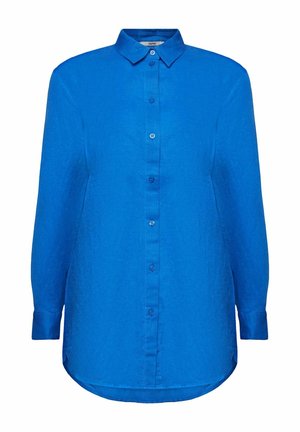 Vrouw die een loszittend blauw overhemd met knopen en opgerolde mouwen en een witte broek draagt, hand in de zak, tegen een effen witte achtergrond.