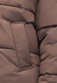 Close-up van een bruin doorgestikt stof met een rechthoekig gestikt zakje op een gewatteerd jack.