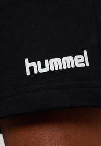 Pantalones cortos deportivos negros con una textura suave y el logo blanco de "hummel" impreso de forma prominente en el dobladillo inferior. Se aprecian detalles de costura.