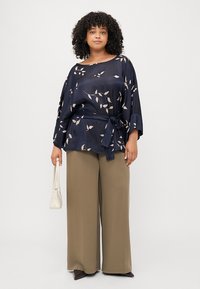 Blusa blu navy con motivo floreale, maniche ampie e cintura in vita, abbinata a pantaloni larghi verdi oliva e una borsa bianca.