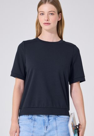 Ung kvinde med langt blondt hår, iført en sort kortærmet skjorte og lyseblå jeans, der holder en bog og solbriller i højre hånd.