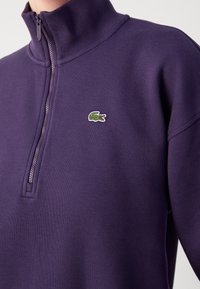 Gros plan sur un pull violet à fermeture éclair sur le quart supérieur, en tissu texturé, avec un petit logo de crocodile vert sur le côté gauche de la poitrine.