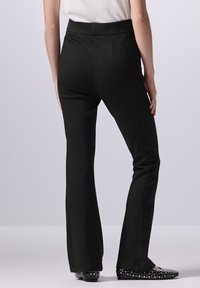 Pantaloni neri a zampa d'elefante con una texture di tessuto liscia e una vita alta, caratterizzati da dettagli di cuciture verticali e indossati con ballerine nere con borchie.