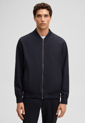 Mężczyzna w czarnej, fakturowanej kurtce na zamek i dopasowanych spodniach, stojący na jednolitym jasnoszarym tle.