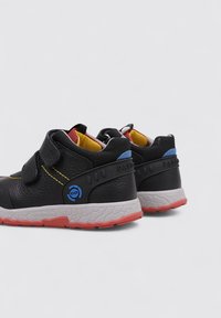 Chaussures noires pour enfants avec une finition texturée, dotées de bandes Velcro, accents rouges et jaunes, et une semelle blanche et rouge avec un motif de semelle.