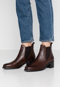 Stivali alla caviglia in pelle marrone con una superficie liscia, pannelli elastici ai lati, un piccolo tab sul retro e un tacco basso a blocco. Abbinati a jeans blu leggermente accorciati.
