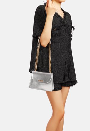 Borsa a tracolla argentata con chiusura a patta, tracolla a catena e accessori in tono oro. Il modello indossa un outfit nero texturizzato.