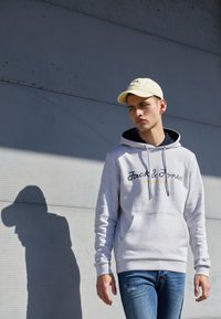 Lys grå hættetrøje med marineblå foring, frontlomme og sort broderet tekst. Kombineret med blå jeans og en lys gul kasket.