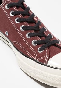 Zapatilla de lona marrón con cordones negros, ojales plateados y una puntera de goma blanca. Presenta costuras blancas y una suela texturizada.