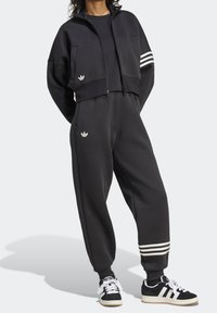 Persona che indossa un completo da ginnastica Adidas nero con le tre strisce bianche sulle maniche e sui polsini dei pantaloni, abbinato a scarpe da ginnastica Adidas nere e bianche.