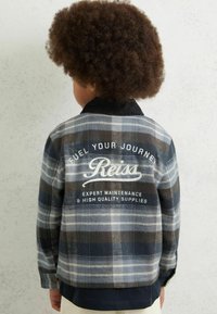 Navy, pilkų ir baltų atspalvių langeliais flanelinė striukė su juodu apykakle. Ant nugaros yra grafika su tekstu ir subtiliu tekstūruotu audiniu.