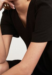 Robe noire à manches courtes avec un décolleté en V, présentant une texture lisse et une coupe décontractée. Teint de peau visible sur les bras découverts.