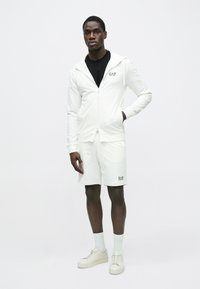 Bela majica s kapuco z zadrgo in ujemajoče kratke hlače iz mehke tkanine. Črn logotip na prsih, skupaj z belimi nogavicami in svetlimi supergami.