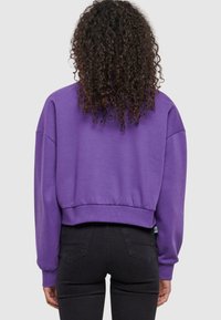 Felpa viola corta in morbido tessuto con maniche ampie e orlo a costine, abbinata a jeans neri.