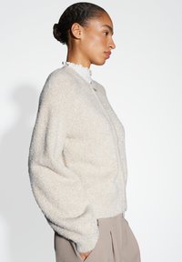 Vrouw met een knot in haar haar, gekleed in een fuzzy crèmekleurige cardigan over een witte gerimpelde coltrui en beige plooibroek, met haar handen in de zakken, met haar gezicht naar rechts.