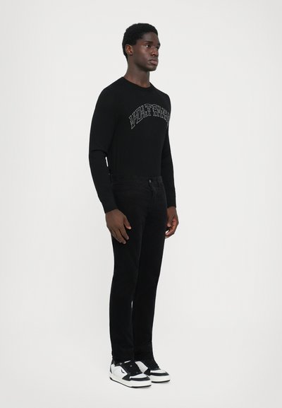 Sort langærmet skjorte med "VOLTAIRE" tryk, parret med sorte kordfløjspants og sort-hvide sneakers. Simple, tætsiddende design.