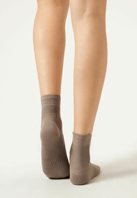 Brown ankle socks made of a soft, textured material with a ribbed cuff and gripper dots on the soles, displayed on bare legs.