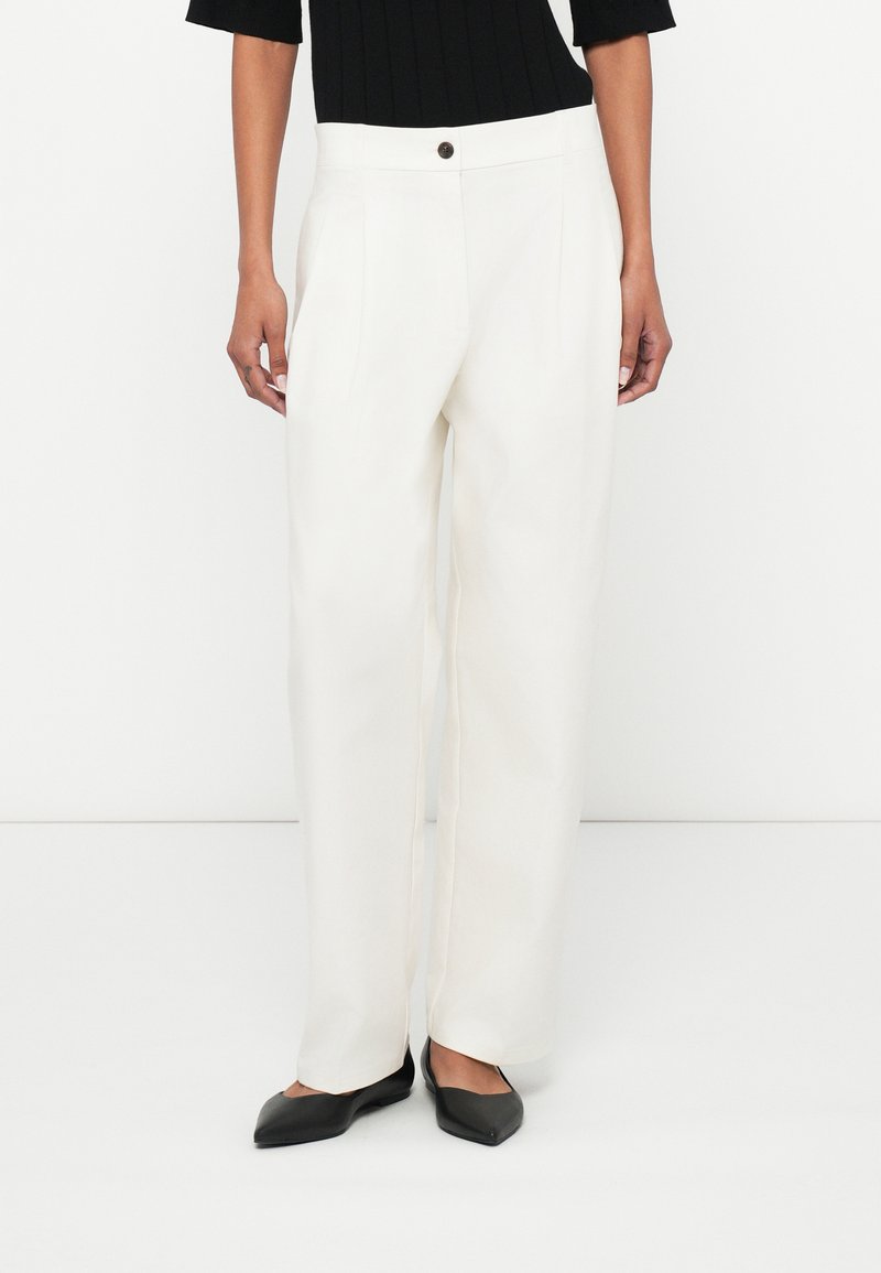 Hvide skræddersyede bukser med høj talje, med folder og enkelt knaplukning. Kombineret med sorte spidse flade sko.