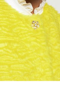 Gul fluffig plagg med en mjuk textur, som har en dekorativ brosch med klara stenar i en blommig arrangemang fram.