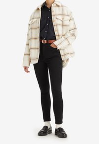 Person wearing a cream plaid button-up jacket over a black shirt tucked into high-waist black pants with a brown belt and black loafers.