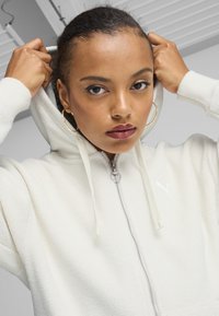 Felpa bianca con zip, texture in pile, cappuccio con cordino e logo ricamato. Modello posizionato con le mani che regolano il cappuccio.