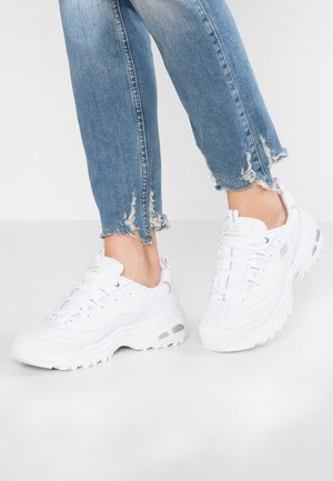 Białe masywne sneakersy noszone z niebieskimi, obcisłymi jeansami z postrzępionym brzegiem, zdjęcie od połowy łydki w dół na jednolitym jasnym tle.
