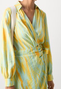 Vietto wrap dress mekansä sininen vaaleassa sävyssä, jossa on keltaisia ja oransseja aaltoilevia kuvioita. Tunne kesäinen kaulus, pitkät hihat ja vyötärölle solmittu yksityiskohta. Pehmeä pinta.