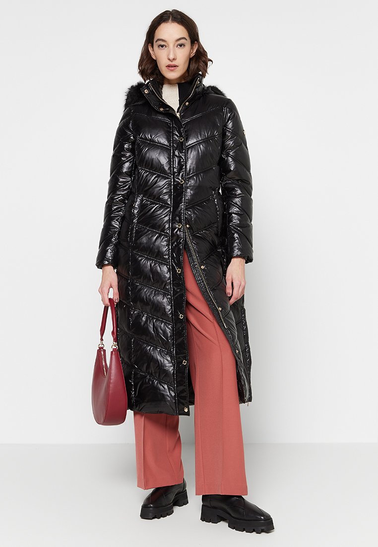 Donna che indossa un lungo cappotto nero trapuntato con cappuccio foderato in pelliccia, pantaloni larghi color ruggine, stivali neri e tiene in mano una borsa rossa.