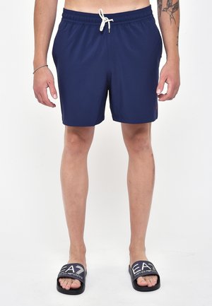 Pantaloni da bagno blu navy con cintura elastica e lacci, dotati di tasche laterali e un piccolo logo. Abbinati a sandali neri in slide.
