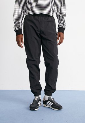 Pantalons de sport noirs à coupe décontractée, avec une taille élastique et un tissu texturé. Associés à des baskets noires avec des accents blancs.