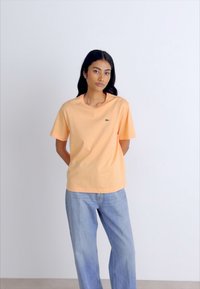 Pfirsichfarbene, kurzärmlige T-Shirt aus Baumwolle mit einem kleinen grünen Logo auf der linken Brust; kombiniert mit hellblauen Jeans.