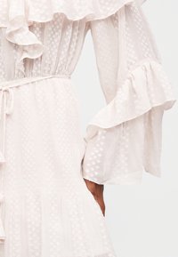 Λευκό υφασμάτινο φόρεμα με φουσκωτά μανίκια, ζώνη στη μέση και διάφανα μακριά μανίκια με ανάγλυφο μοτίβο από μικρές τελείες.