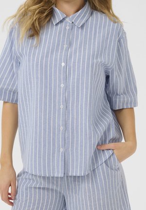 Kobieta w jasnoniebieskiej koszuli z krótkim rękawem zapinanej na guziki oraz dopasowanych spodniach z pionowymi białymi paskami, jedna ręka w kieszeni.