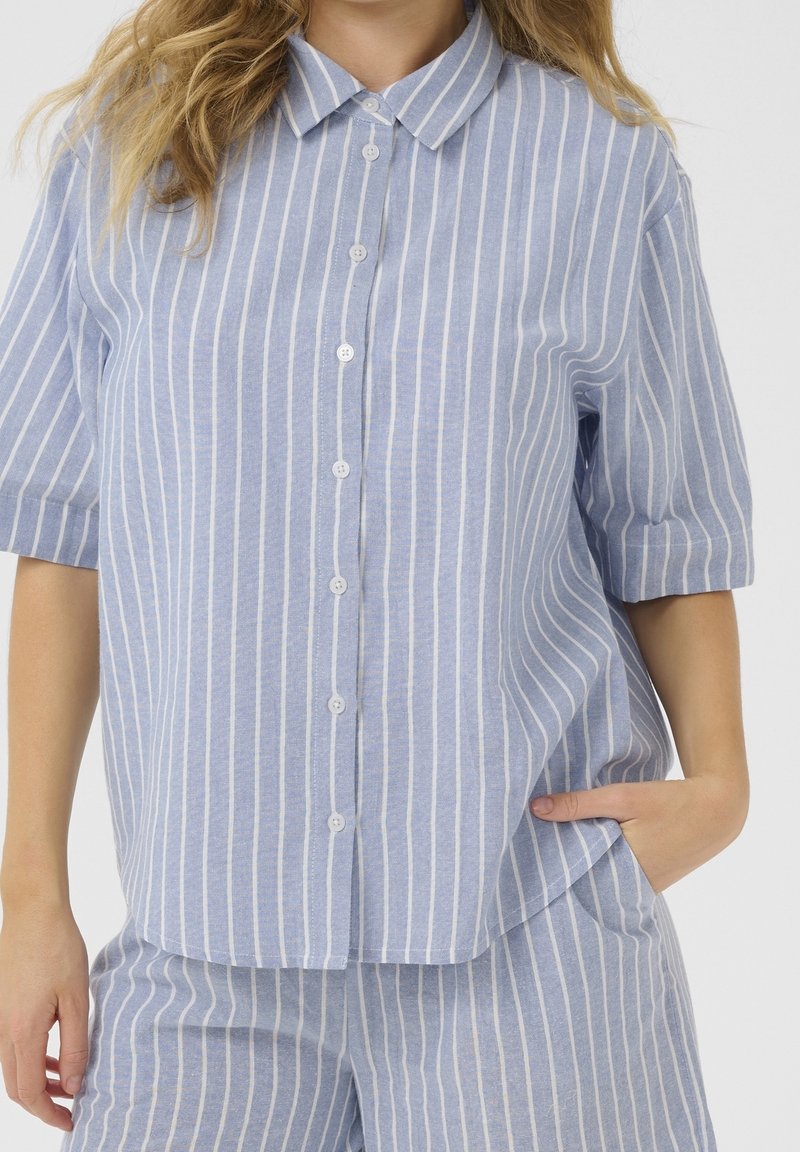 Femme portant une chemise bleu clair à manches courtes boutonnée et un pantalon assorti à rayures blanches verticales, une main dans la poche.