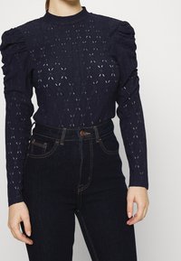 Tmavě modrý svetr s dlouhým rukávem a vysokým límcem, s nařasenými rameny, diamantovými vzory a texturovanou pleteninou. V kombinaci s tmavými džíny.