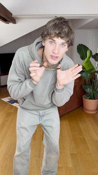 Grå sweatshirt med hætte, lysegrå jeans og en perlekæde. Indendørs setting med en potteplante i baggrunden.