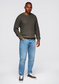 Suéter de punto verde oscuro con puños acanalados, combinado con jeans azul claro y zapatillas grises. El modelo está de pie sobre un fondo liso.