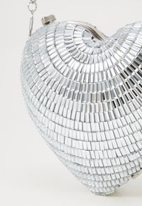Clutch en forme de cœur fabriqué à partir de carreaux en miroir argentés, avec un fermoir en métal et une bandoulière en chaîne. Texture lisse avec surface réfléchissante.