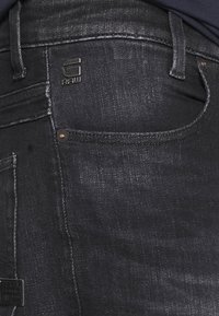 Mörka denimjeans med en slät textur, minimal blekning och en detalj med svart metall-logotyp. Inkluderar fem fickor och kopparnitar.