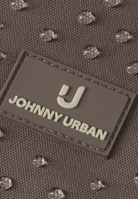 Detailný záber na hnedú látku s kvapkami vody a gumovou nášivkou s logom "Johnny Urban" a štylizovaným písmenom J.