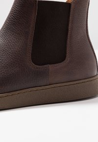 Botín de cuero marrón con acabado texturizado, paneles laterales elásticos y suela de goma marrón. Presenta una costura a lo largo de la base.