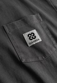 Temno sivo platneno žep z belim kvadratnim obližem, na katerem je črno geometrijsko logotip in pod njim napis "GABBA".