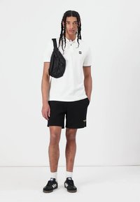 Hvit polo skjorte, svarte shorts, svart skulderveske. Modellen har på seg svarte sneakers med hvite detaljer, hvite sokker, og har fletninger.