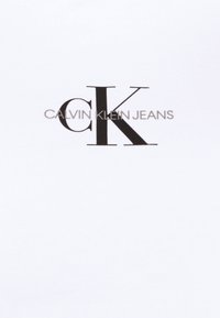 T-shirt in cotone bianco con un prominente logo "CK" nero e il testo "CALVIN KLEIN JEANS" sottostante, stampato in un moderno carattere sans-serif.