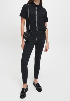 Czarna koszula z krótkim rękawem z logo na środku, w połączeniu z ciemnymi obcisłymi jeansami i błyszczącymi czarnymi mokasynami. Dodatki to mała torebka na ramię.