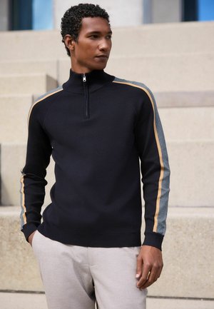 Man wearing a black zip-up sweater with gray and beige stripes on the sleeves, standing near light-colored concrete steps.