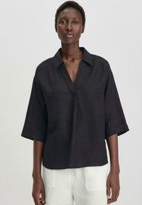 Camicia nera in lino a maniche corte con collo a camicia e scollatura a V frontale. Il tessuto ha una texture sottile ed è di vestibilità rilassata.